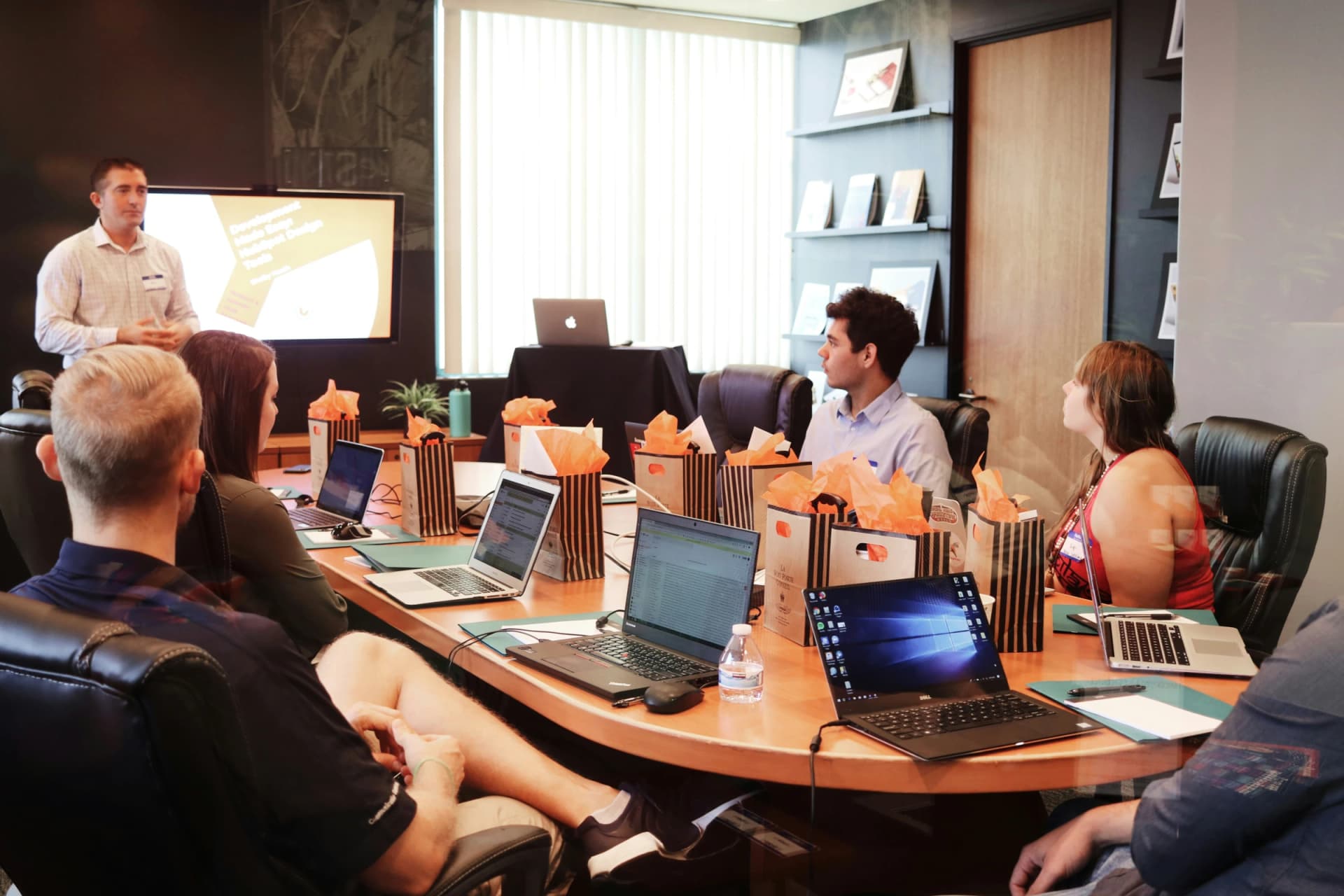 Man presenting in front of a wall screen to a group seated with laptops around a conference table set with gift bags