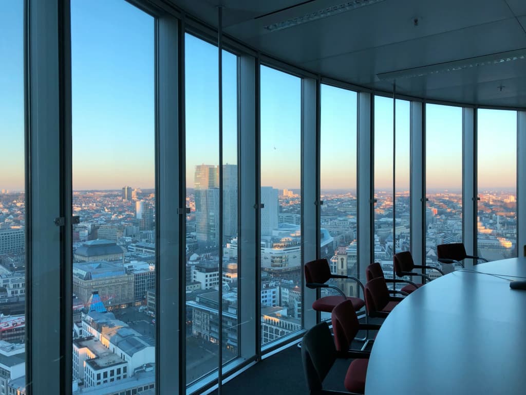 Empty conference room with red chairs around a curved table, behind floor-to-ceiling windows overlooking a city skyline at sunset