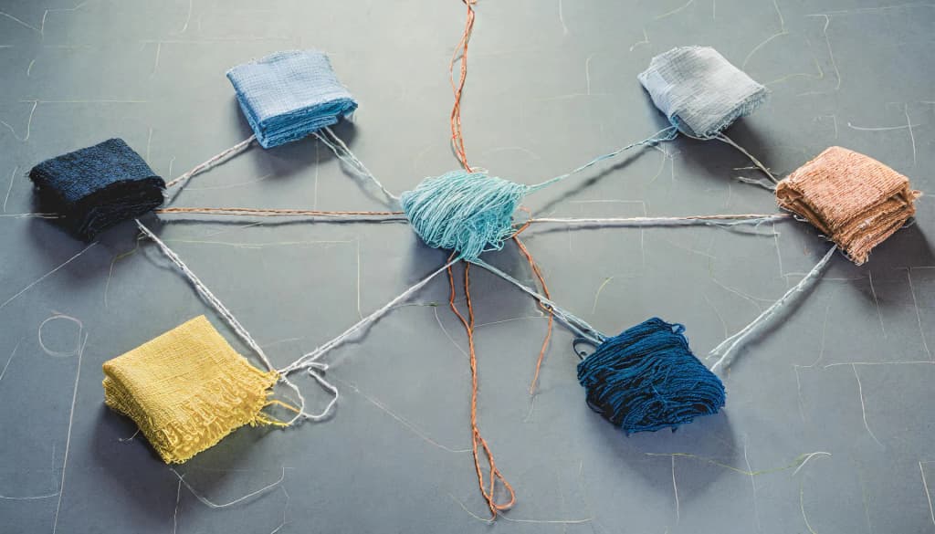Folded fabric swatches in yellow, navy, light blue, gray, and copper, linked by threads to a tangled ball of turquoise yarn in the center like a network